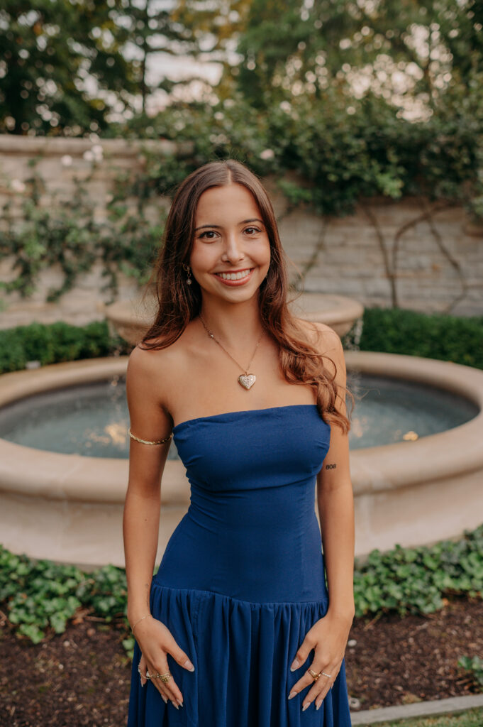 High school senior girl in a navy blue dress smiling in front of a stone garden fountain — personalized senior portrait location near Barrington IL